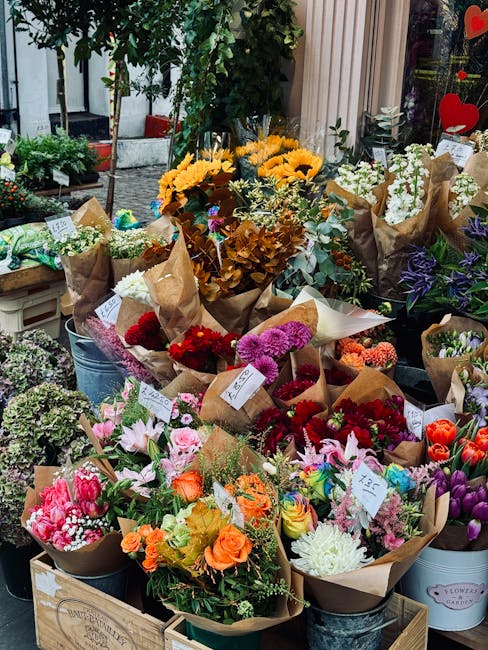 An outdoor flower shop display featuring a variety of fresh floral arrangements and bouquets. Prominent in the foreground are bouquets wrapped in brown kraft paper with vibrant pink, orange, white, an