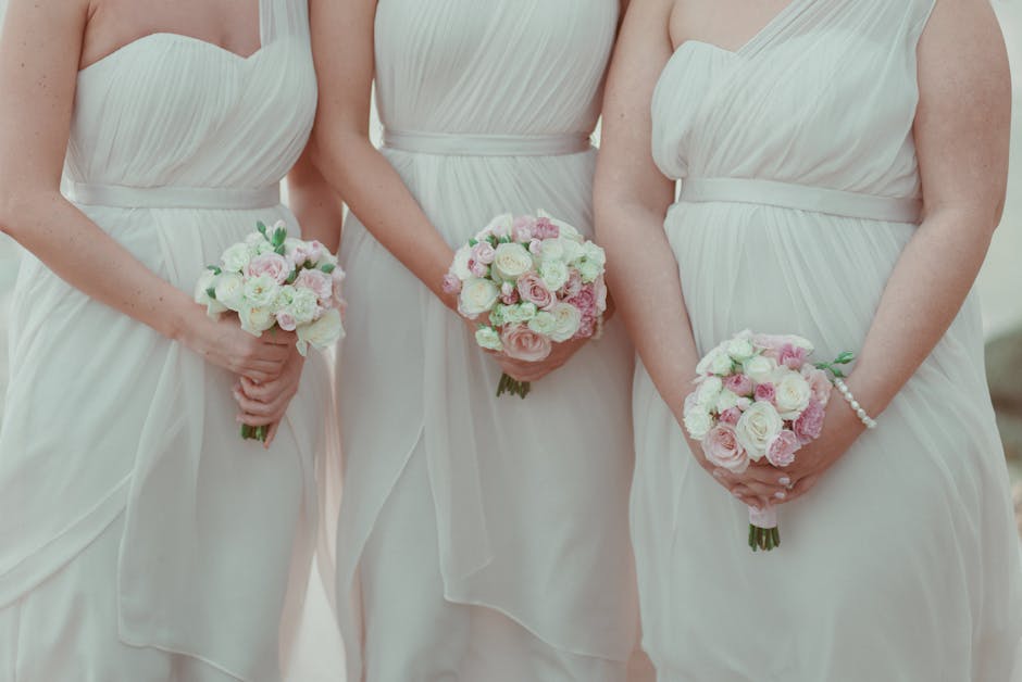 Three women in elegant white bridesmaid dresses standing side by side, each holding a round bouquet arrangement of fresh, soft pink and white roses, peonies, and ranunculus, accented with green foliag