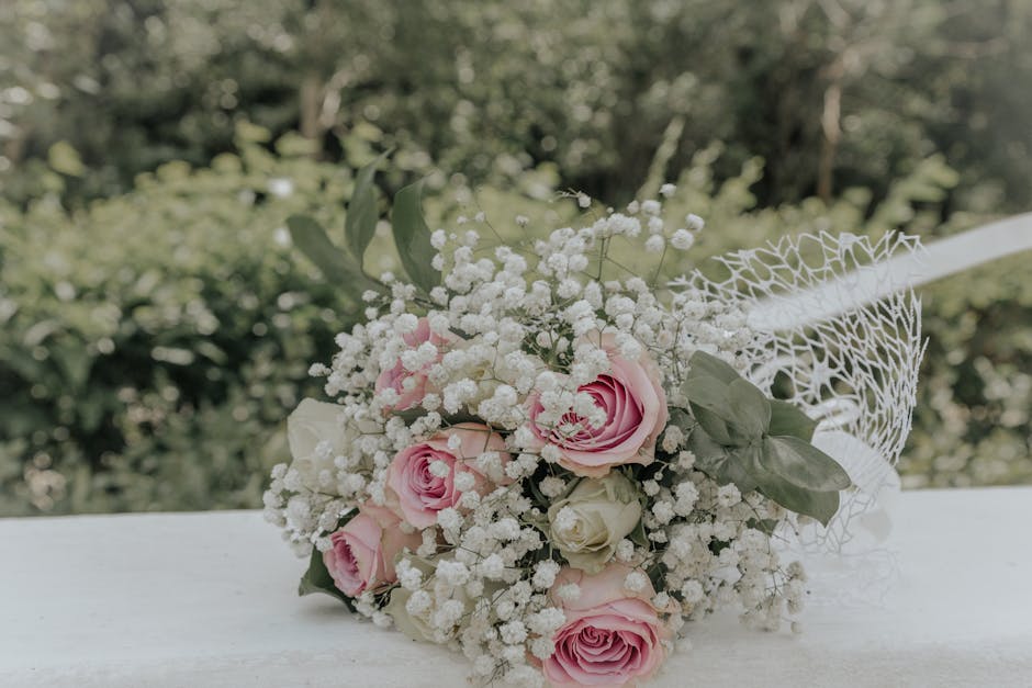 A delicate floral bouquet featuring soft pink roses and white roses, complemented by sprigs of baby's breath, arranged with fresh green foliage, and wrapped in white netting material. The arrangement 