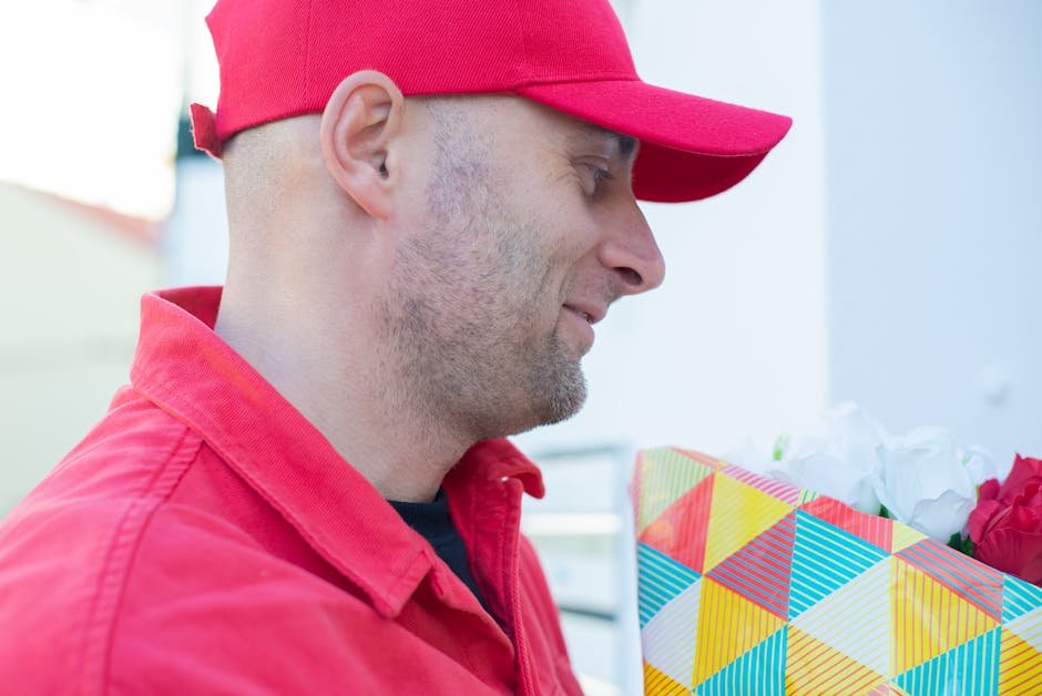 A delivery person in a red cap and matching red jacket is holding a floral gift package wrapped in colorful, geometric-patterned paper, with white tissue paper and red roses visible inside. The person