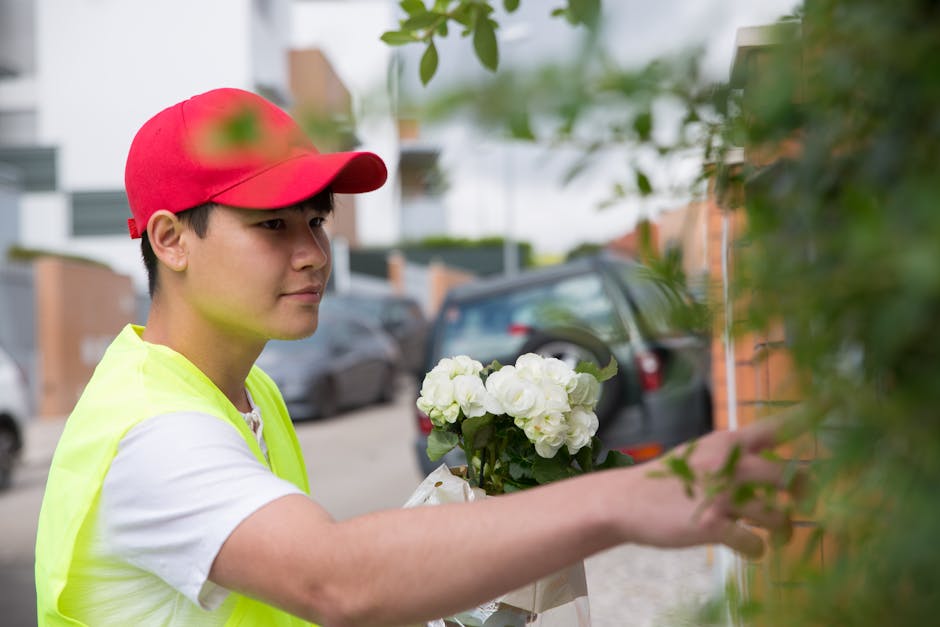 A young male florist wearing a red cap and a yellow vest is placing a bouquet of fresh white hydrangeas and roses against a leafy green wall outdoors. The bouquet is wrapped in white paper and kept ne