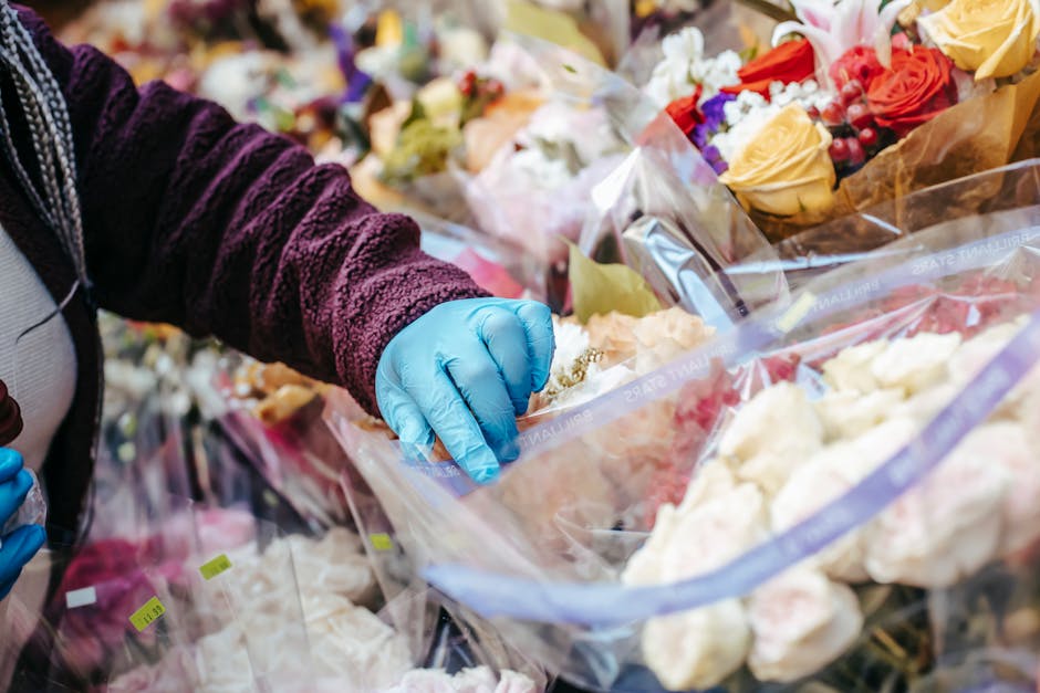A person wearing a dark purple textured jacket and blue gloves is selecting a floral arrangement from a display of various bouquets wrapped in clear and colored plastic at South Kensington Florist. Th
