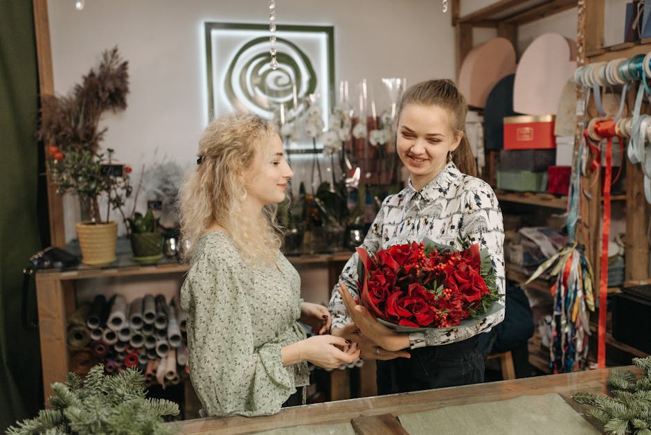A young woman with light brown hair tied back, wearing a patterned shirt, is handing a large bouquet of vibrant red roses, wrapped with green foliage and decorative paper, to a woman with curly blonde