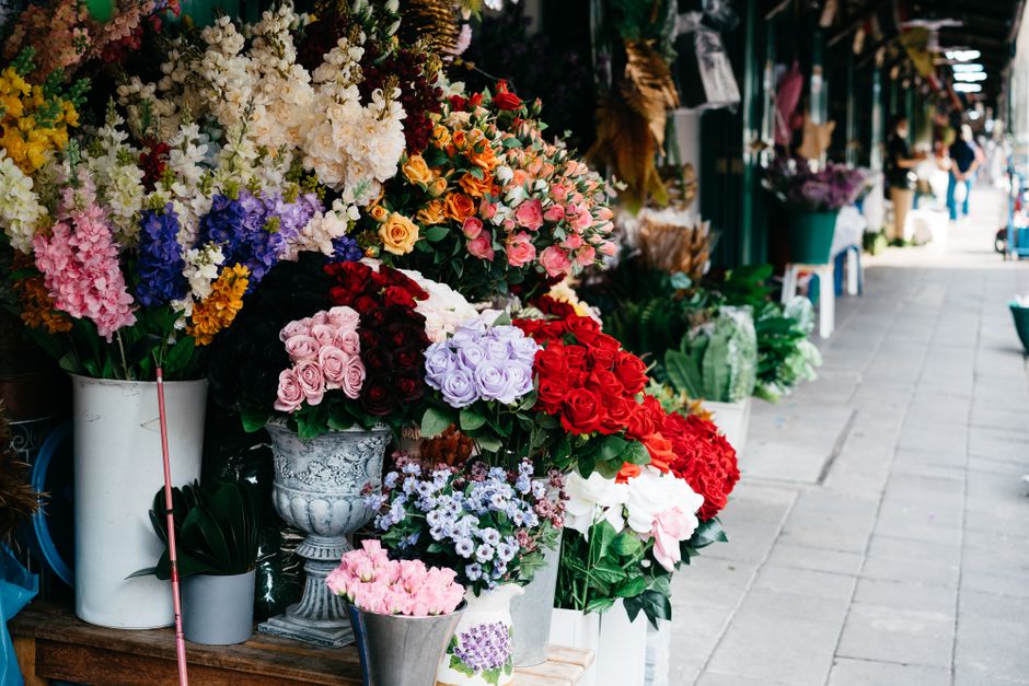 A vibrant outdoor flower shop display featuring an abundance of fresh floral arrangements in various colors and styles. The arrangement includes bouquets of roses in shades of pink, red, and lavender,