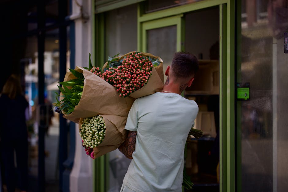 A person with short dark hair and a white t-shirt is seen from behind, carrying a large bouquet of fresh flowers wrapped in brown paper. The bouquet includes pink berries, white blossoms, and assorted